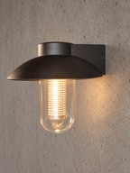 A matte black outdoor wall light fixture with a wide circular shade and a clear glass bulb, mounted on a textured concrete wall and casting a warm glow.