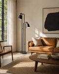 A modern black dual-arm floor lamp stands beside a tan leather sofa in a sunlit living room with a wood coffee table, woven rug, and large abstract wall art.