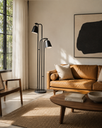 A modern black dual-arm floor lamp stands beside a tan leather sofa in a sunlit living room with a wood coffee table, woven rug, and large abstract wall art.