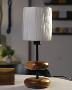 A serene forest retreat bedside features the 'Earthy Wood Glow' table lamp—teak wood base, sleek metal stem, and off-white fabric shade—on a rustic wooden table, with a golden glow illuminating books, a ceramic mug, and a lush forest view through a window