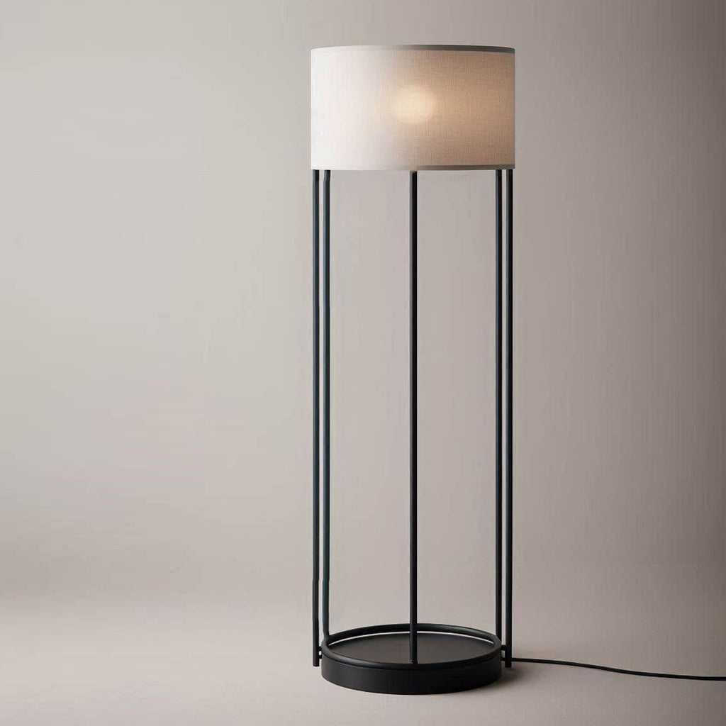 A sophisticated floor lamp with a minimalist black metal frame and an elegant cylindrical beige shade. The lamp is switched on, emitting a warm glow, and stands against a neutral background.