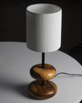 A warm teak wood table lamp with a sleek metal stem and soft off-white fabric shade glows gently on a rustic wooden bedside table in a forest retreat bedroom, surrounded by nature-inspired decor and a view of pine trees through a window.