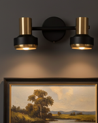 Wall-mounted light fixture with two gold bulbs on a dark wall above a framed landscape painting.