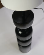 Contemporary table lamp featuring a glossy black geometric base and a white cylindrical lampshade on a desk.