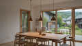 Dining area with wooden table and chairs in a room with large windows showing a view of a house and trees.