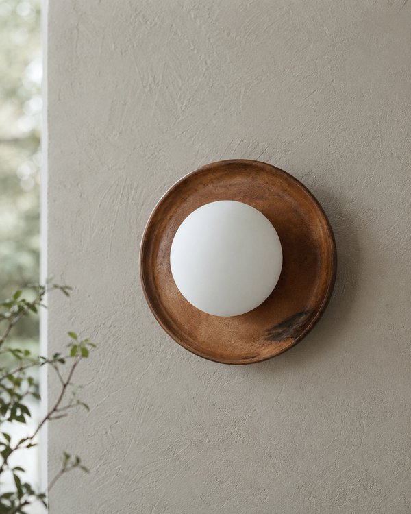 Handcrafted solid wood wall sconce featuring an 8-inch natural wooden backplate with a centered 4-inch frosted opal glass globe, photographed in soft daylight against a textured plaster wall in a warm mid-century modern interior.