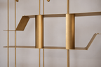 A close-up of a modern brass shelving unit with minimalist geometric metal plates and vertical rods against a neutral light gray background.