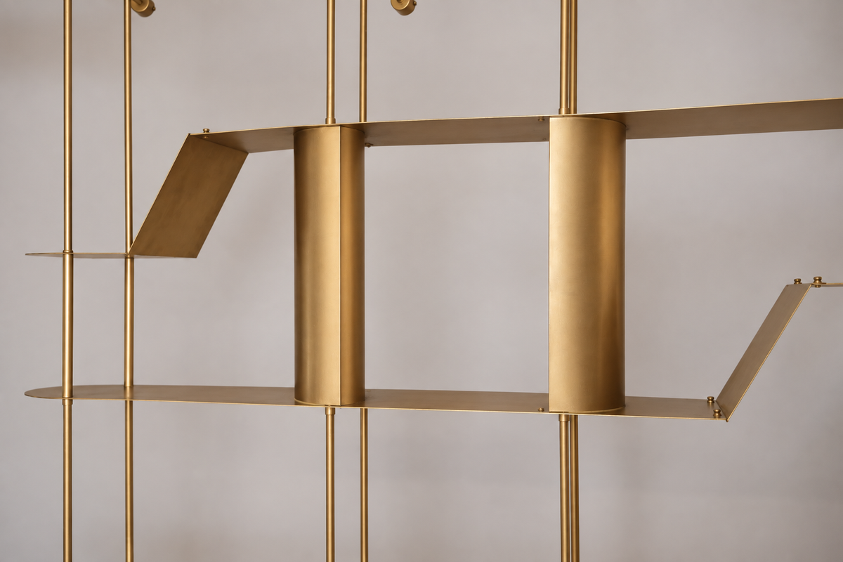 A close-up of a modern brass shelving unit with minimalist geometric metal plates and vertical rods against a neutral light gray background.