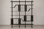 A modern black metal shelving unit with cylindrical and stepped open shelves, shown against a neutral beige wall and concrete floor.
