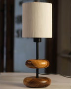 On a honey-toned wooden bedside table in a forest retreat, the 'Earthy Wood Glow' lamp with its teak wood base, metal stem, and off-white shade casts a soft light, framed by a moss-green wall, nature sketches, and a view of tall pine trees outside.