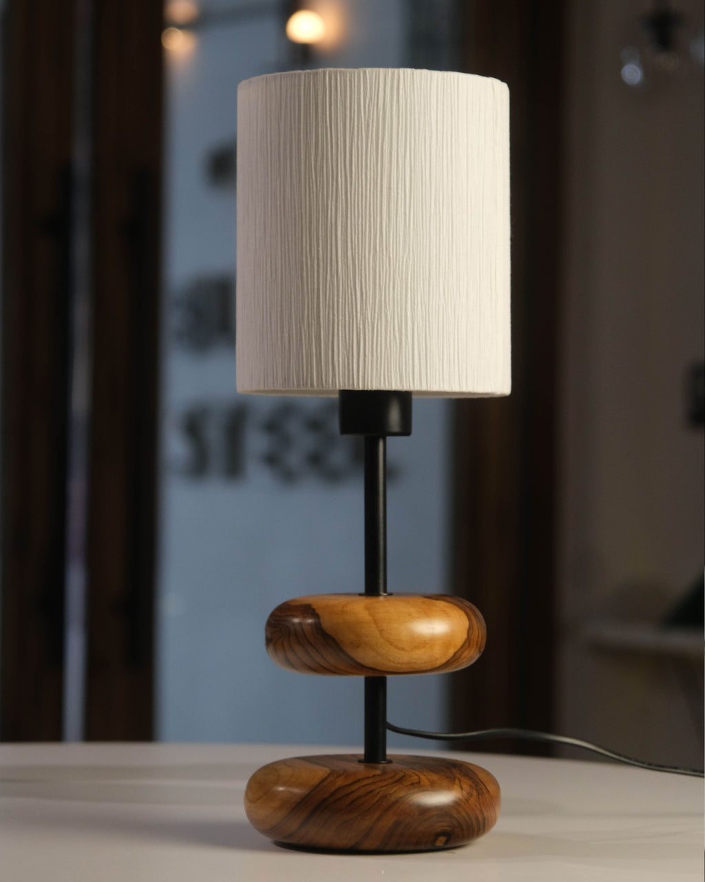 The 'Earthy Wood Glow' lamp, featuring a rich teak wood base, neutral metal stem, and off-white fabric shade, sits on a reclaimed wood bedside table in a cozy forest retreat, with books and lavender nearby, illuminated against a woodland backdrop