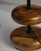 The 'Earthy Wood Glow' lamp, with its earthy teak wood base, minimalist metal stem, and soft off-white shade, rests on a wooden bedside table in a tranquil forest retreat, surrounded by natural decor and a scenic view of pine trees and dappled sunlight.