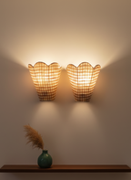 Two wall-mounted lamps with checkered shades on a plain wall, with a green vase and dried plants on a shelf below.