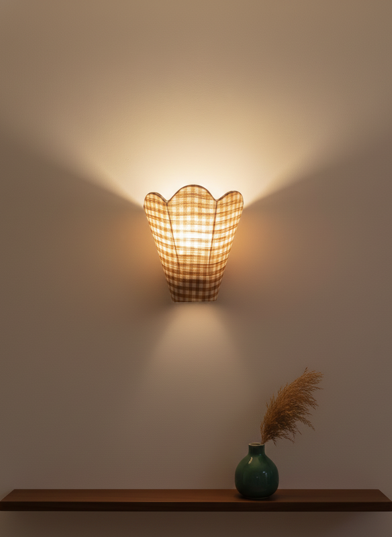 Wall-mounted lamp with a checkered pattern casting light on a plain wall, with a vase and dried plants on a shelf below.