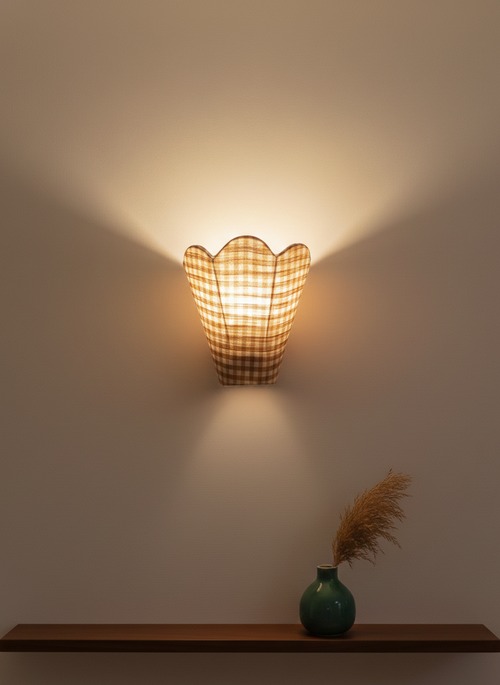 Wall-mounted lamp with a checkered pattern casting light on a plain wall, with a vase and dried plants on a shelf below.
