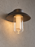 A modern outdoor wall sconce with a dark bronze metal frame and a clear glass cylinder, glowing with a warm yellow light against a textured concrete wall.