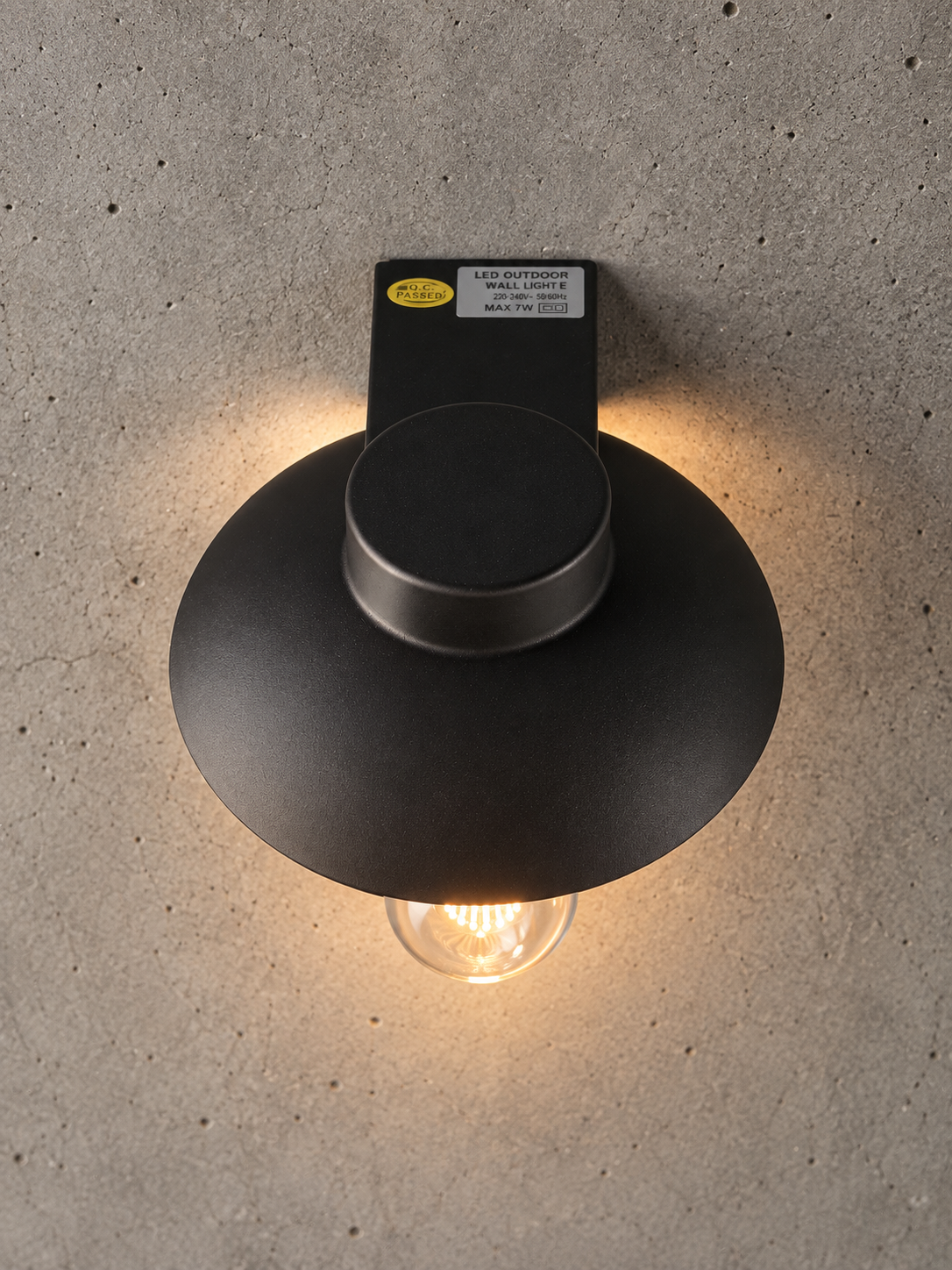 A matte black LED outdoor wall light fixture with a wide circular shade and a clear glass bulb, mounted on a textured concrete wall and casting a warm glow.